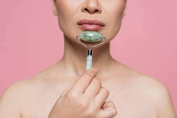Cropped view of woman with naked shoulders massaging chin with jade roller isolated on pink  - Photo, Image