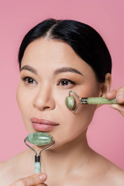 Asian woman with naked shoulders massaging face with jade rollers isolated on pink  - Foto, imagen