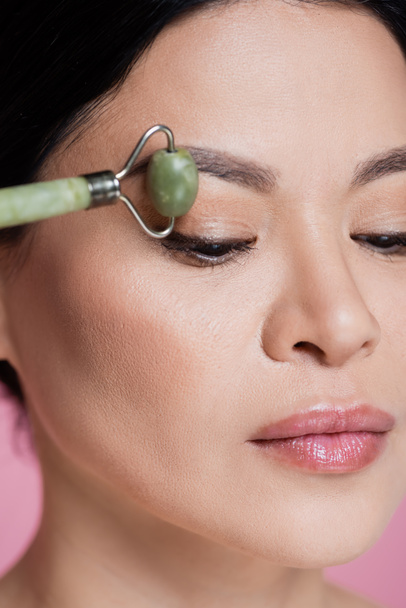 Close up vista da mulher asiática massageando rosto com rolo de jade desfocado isolado em rosa  - Foto, Imagem