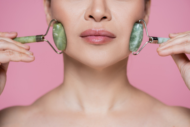 Cropped view of woman massaging cheeks with jade rollers isolated on pink  - Fotó, kép