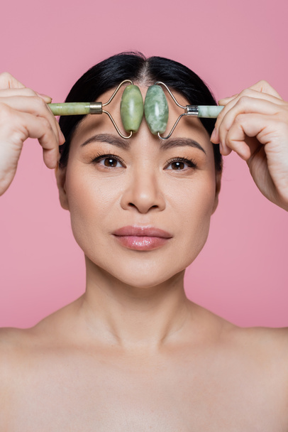 Asian woman with naked shoulders massaging forehead with jade rollers isolated on pink  - Valokuva, kuva
