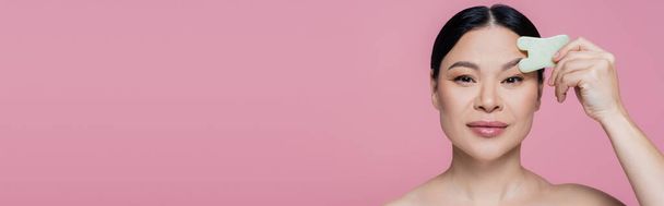 Mulher asiática com ombro nu massageando rosto com pedra de jade isolado em rosa, banner  - Foto, Imagem