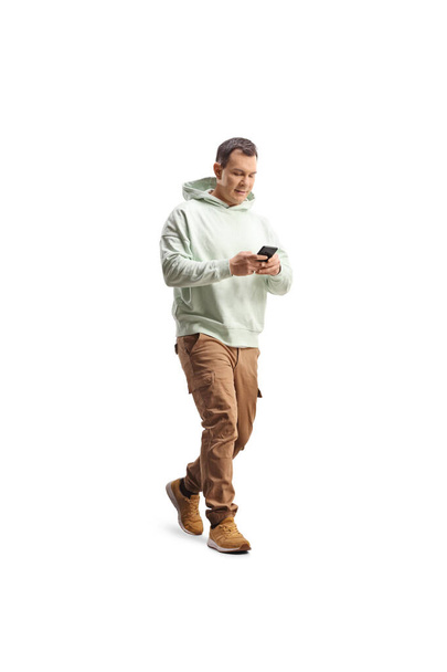 Full length shot of a casual man walking and typing on a smartphone isolated on white background - 写真・画像