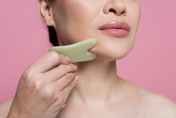 Cropped view of woman with naked shoulder massaging chin with jade stone isolated on pink  - Foto, afbeelding