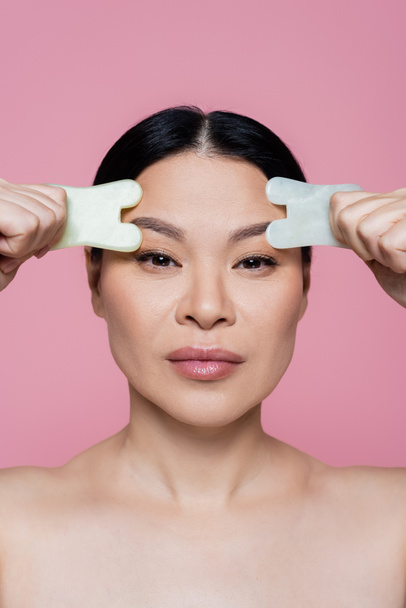 Asian woman with naked shoulders holding jade stones near eyebrows isolated on pink  - Photo, Image