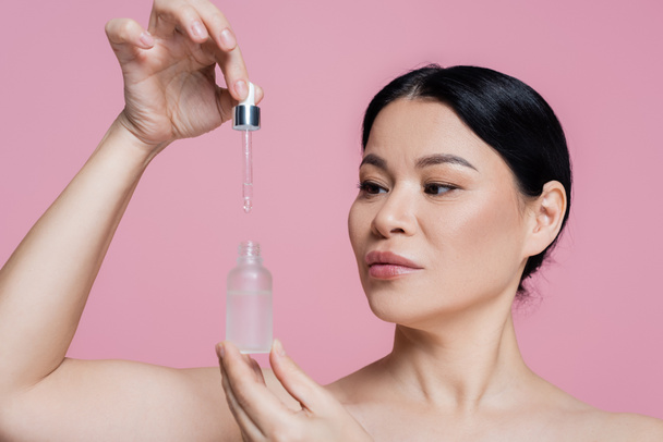 Asian woman with naked shoulders holding dropper and serum isolated on pink  - Φωτογραφία, εικόνα