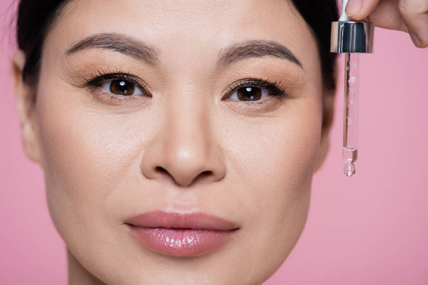 Close up view of asian woman looking at camera and holding dropper with serum isolated on pink  - Fotó, kép