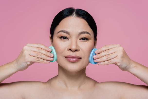 Asian woman with naked shoulders cleaning face with sponges isolated on pink  - Foto, imagen