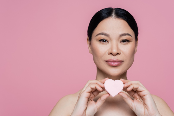 Asian woman with naked shoulders holding heart shaped beauty blender isolated on pink  - Foto, Imagen