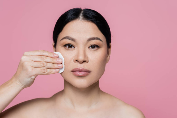 Asian woman with naked shoulders cleaning face with cotton pad isolated on pink  - Foto, immagini