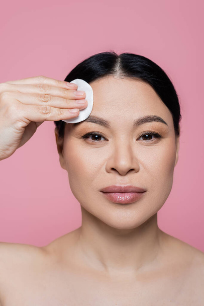 Asian woman looking at camera while cleaning face with cotton pad isolated on pink  - Photo, Image