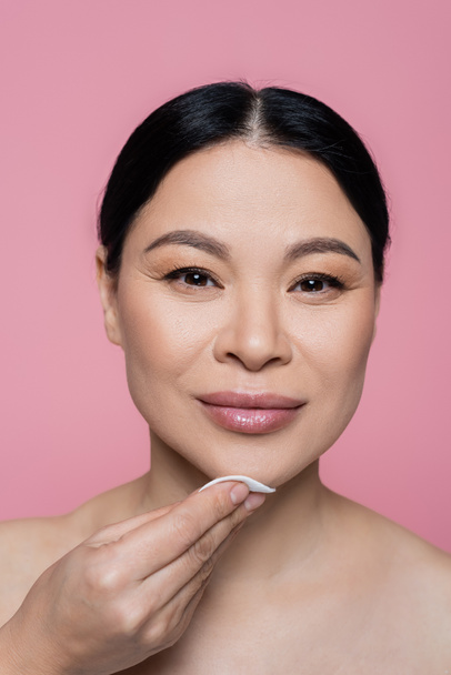 Asian woman looking at camera while cleaning face with cotton pad isolated on pink  - Fotografie, Obrázek