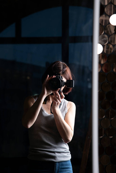 A middle-aged woman photographer looks at herself in the mirror in the semi-darkness of a photo studio. - Фото, изображение