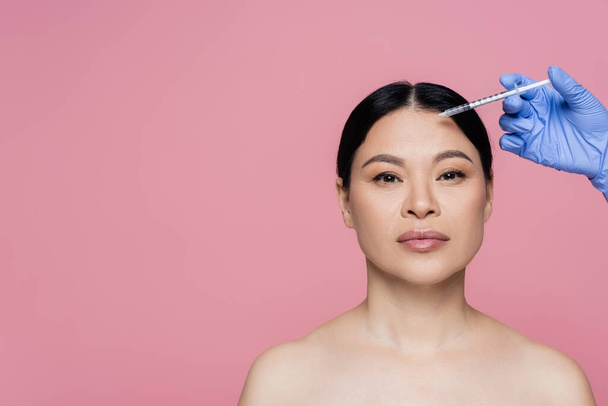 Kosmetikerin in Latex-Handschuh hält Spritze in Stirnnähe asiatischer Frau isoliert auf rosa  - Foto, Bild
