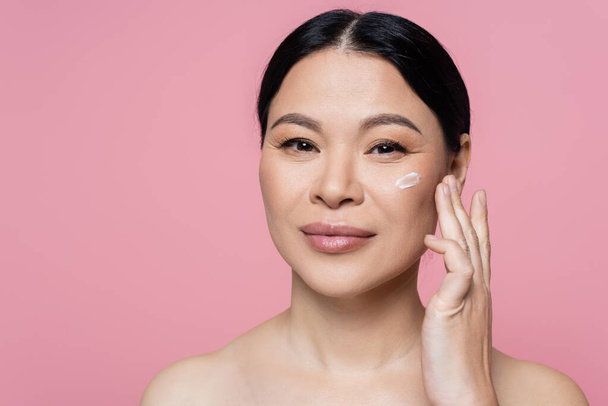 Asian woman applying cosmetic cream and looking at camera isolated on pink  - Fotoğraf, Görsel