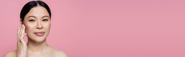Asian woman with naked shoulders applying cosmetic cream and looking at camera isolated on pink, banner  - Foto, Imagem