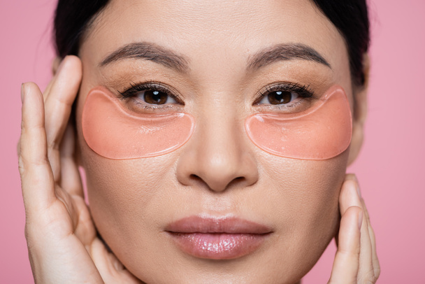 Vue rapprochée de la femme asiatique avec des patchs touchant le visage isolé sur rose  - Photo, image