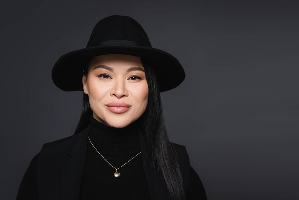 Portrait of stylish asian woman in fedora hat looking at camera isolated on dark grey - Fotografie, Obrázek
