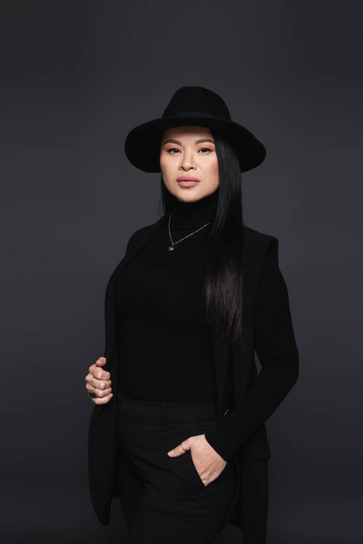 Stylish asian woman in fedora hat posing and looking at camera isolated on dark grey - Photo, Image