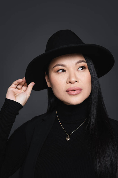 Portrait of stylish asian woman adjusting fedora hat isolated on dark grey - Foto, Bild