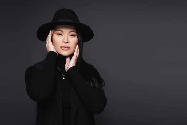 Stylish asian woman in fedora hat touching face isolated on dark grey - Foto, imagen