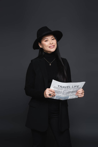 Cheerful asian woman in fedora hat and jacket holding newspaper isolated on dark grey - 写真・画像
