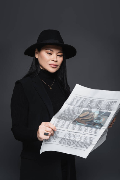 Asiatin mit Fedora-Hut und Blazer liest Zeitung isoliert auf dunkelgrau - Foto, Bild