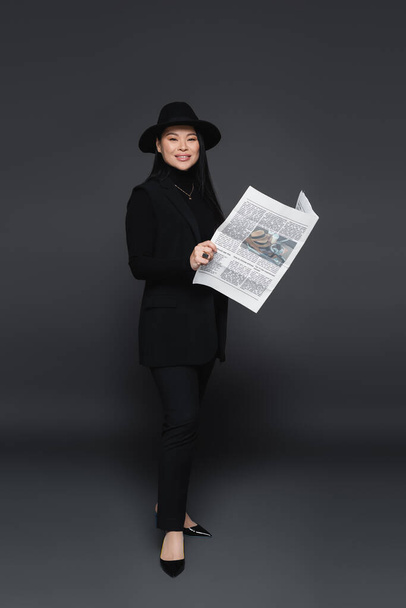 Full length of smiling asian model in fedora hat holding newspaper on dark grey background  - Photo, Image