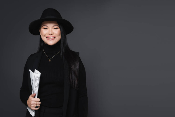Smiling asian woman in jacket and fedora hat holding newspaper isolated on dark grey - Photo, Image