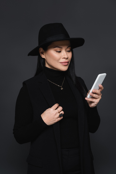 Femme asiatique élégante en chapeau fedora en utilisant un smartphone isolé sur gris foncé - Photo, image