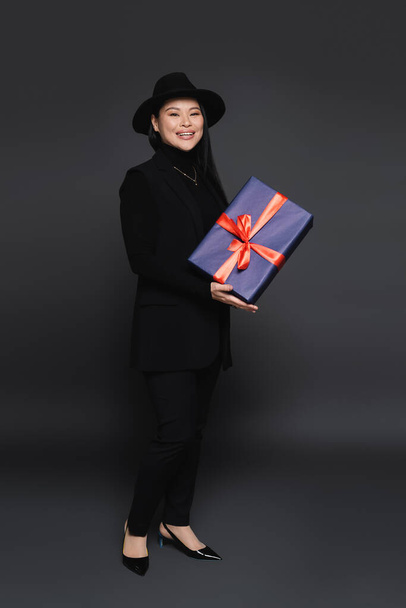 Full length of smiling asian woman in fedora hat holding gift on dark grey background  - Foto, afbeelding