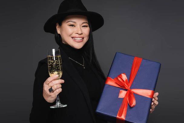 Cheerful asian woman in blazer and fedora hat holding champagne and present isolated on dark grey - Foto, Imagem