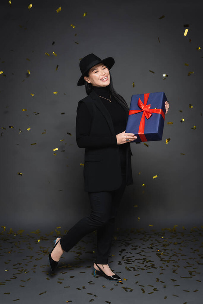Full length of positive asian woman in fedora hat holding gift under falling confetti on dark grey background  - 写真・画像