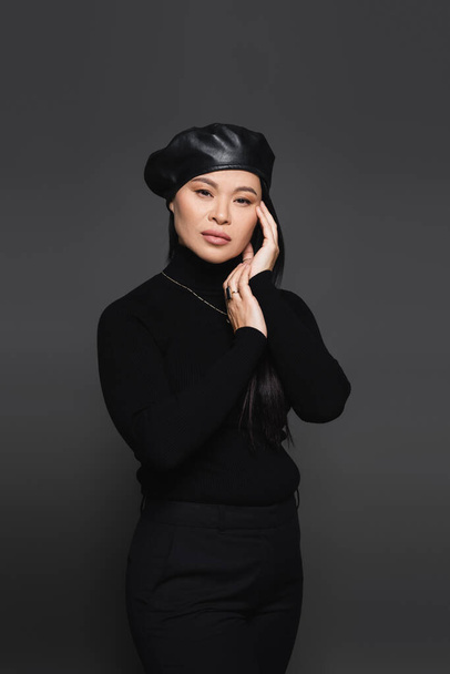 Stylish asian woman in beret and turtleneck touching face isolated on dark grey - Foto, Bild
