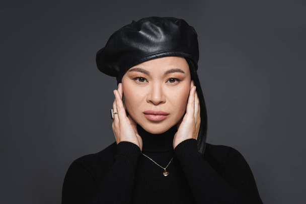 Portrait of stylish asian woman in beret and turtleneck touching face isolated on dark grey - Photo, Image
