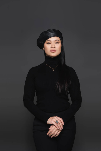 Fashionable asian woman in beret posing and looking at camera isolated on dark grey - Фото, изображение