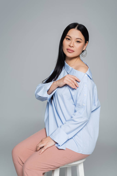 Asian woman in blouse with naked shoulder sitting on chair isolated on grey  - Fotografie, Obrázek