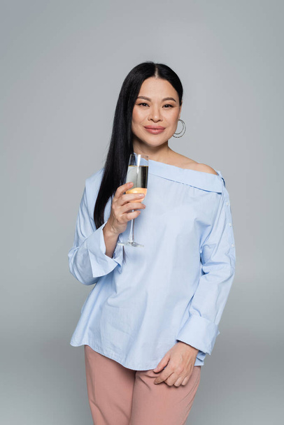 Elegante donna asiatica in possesso di un bicchiere di champagne e sorridente isolato sul grigio  - Foto, immagini