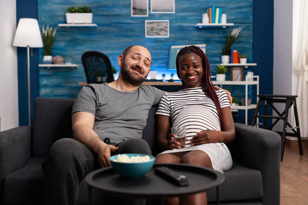Feliz pareja multirracial disfrutando de la televisión y esperando al bebé en casa - Foto, imagen