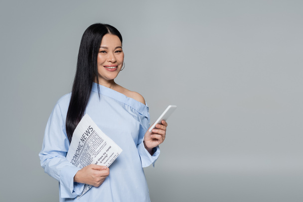 Smiling asian woman in blouse holding cellphone and economic newspaper isolated on grey  - Photo, Image