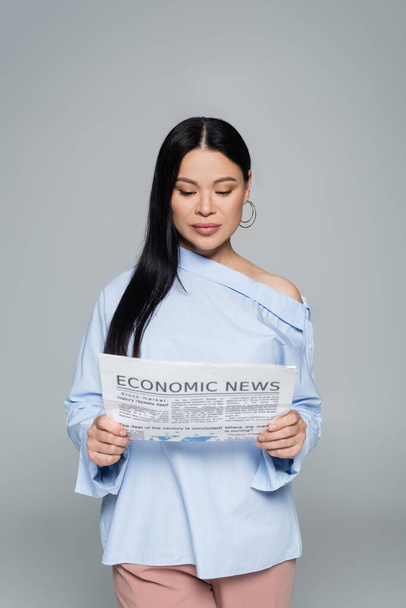 スタイリッシュなブルネットアジアの女性はグレーで隔離された経済ニュースを読んで  - 写真・画像