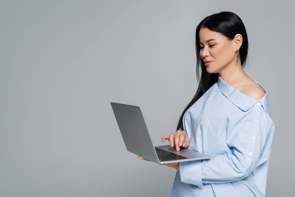 Азиатка в блузке с голым плечом с ноутбуком, изолированным на сером  - Фото, изображение