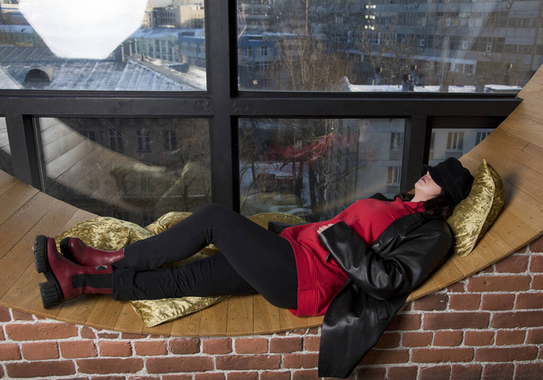 Una adolescente con una chaqueta de cuero está acostada en un gran alféizar junto a una gran ventana redonda. - Foto, imagen