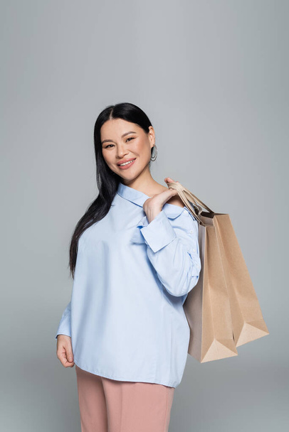 Elegante mulher asiática segurando sacos de compras e olhando para a câmera isolada no cinza  - Foto, Imagem