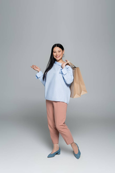 Positive asian model holding shopping bags and pointing with finger on grey background  - Zdjęcie, obraz