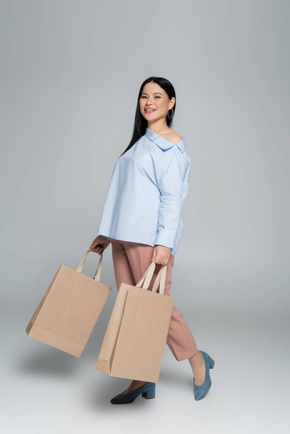 Smiling asian shopaholic holding shopping bags on grey background  - Foto, afbeelding
