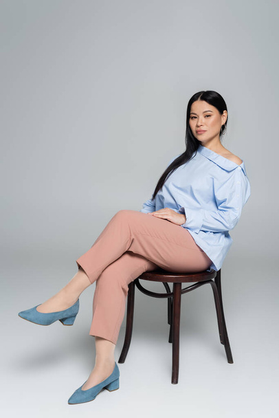 Stylish asian woman positing on chair and looking at camera on grey background  - Foto, immagini