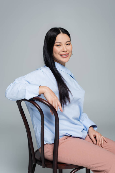 Positive asian woman in blouse sitting on chair and looking at camera isolated on grey  - Фото, зображення