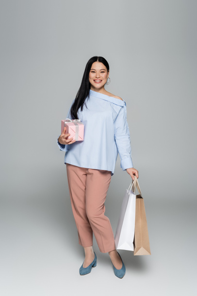 Cheerful asian woman holding gift and shopping bags on grey background  - Valokuva, kuva