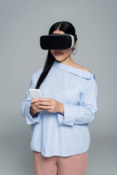 Стильная женщина в VR-гарнитуре с мобильным телефоном изолирована на сером  - Фото, изображение